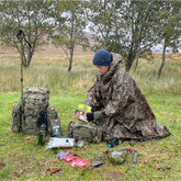 Person wearing a DD Hammocks poncho tarp, surrounded by outdoor gear and backpacks in a natural setting.